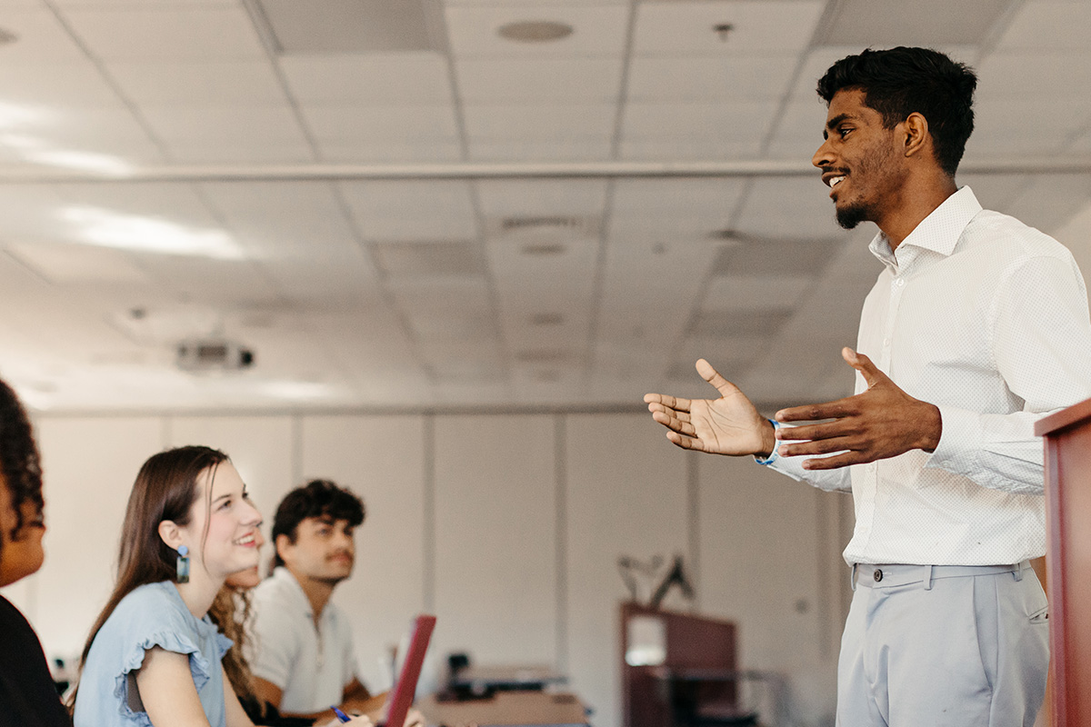 Male student in white shirt and grey slacks stands before classroom and speaks.