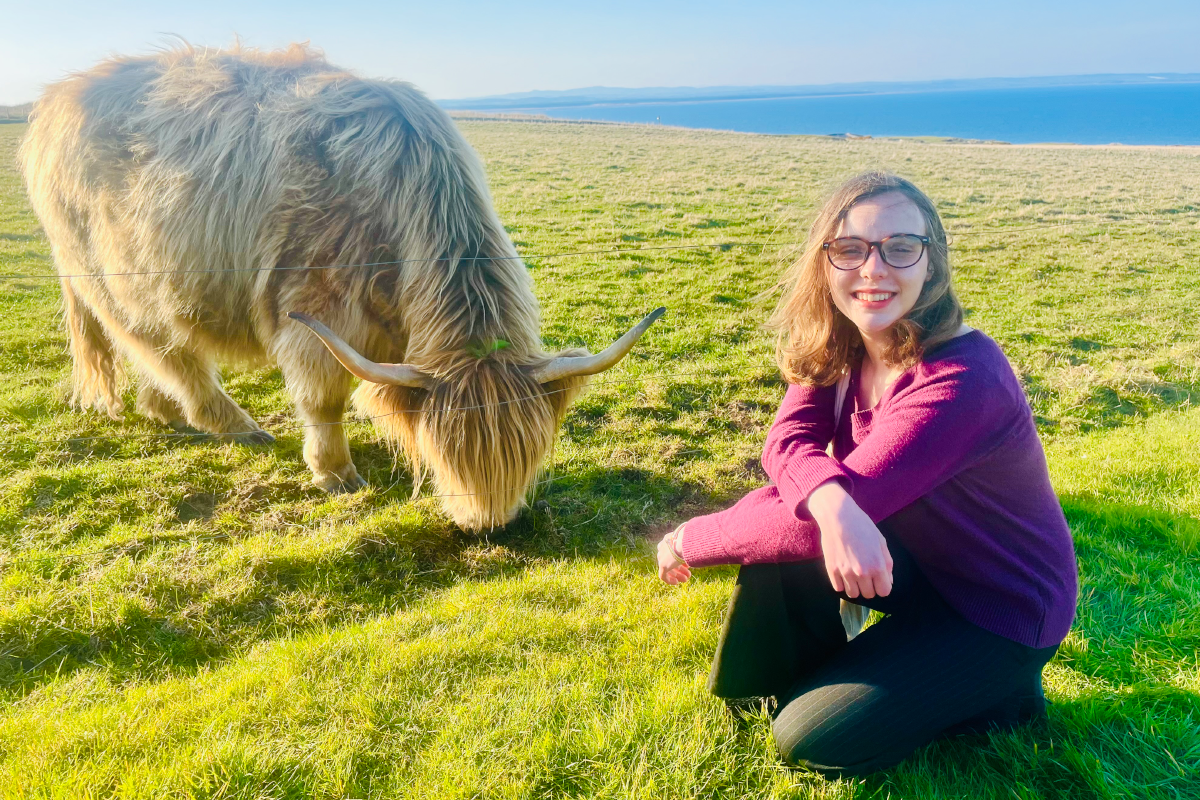 Studying abroad at St. Andrews. Sarah sits in front of a highland cow on the rolling hills of Scotland.